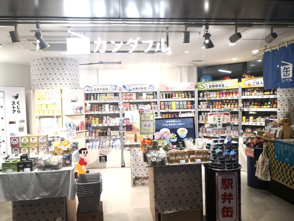 （カンダフル店舗写真）
新橋駅構内にある缶詰め専門店「カンダフル」の店内写真。天井には大きく「カンダフル」のロゴサイン。お魚の缶詰、長期保存食、駅弁缶など多種多様な缶詰めが棚にずらりと陳列されている。左側には「くじらストア」のコーナーも見える。「缶（カン）」＋「ワンダフル（Wonderful）」を組み合わせた造語ネーミングの実例として撮影。